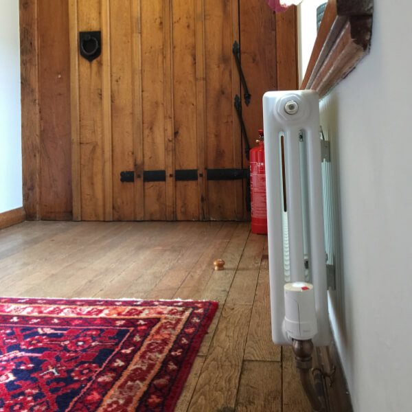 A photo of a hallway in Bossington Hall Hotel, with a wooden door in the middle of the image and a rug on the wooden floor. There is a cast iron radiator on the wall beneath the window and a Wireless Radiator Valve fitted.
