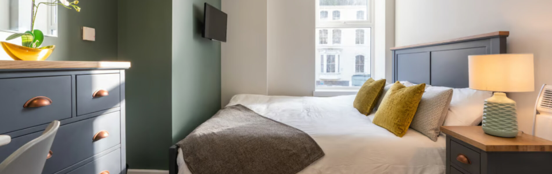A photo of a bedroom in a hotel with a double bed and bedside lamp. There is an Electric Switch on the wall to control the heating.